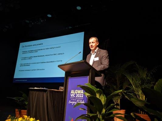 Man in suit stands behind a lectern with a screen behind him